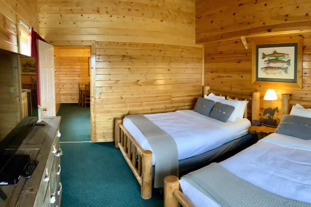 A wooden cabin room with two beds, a desk, and a lamp.