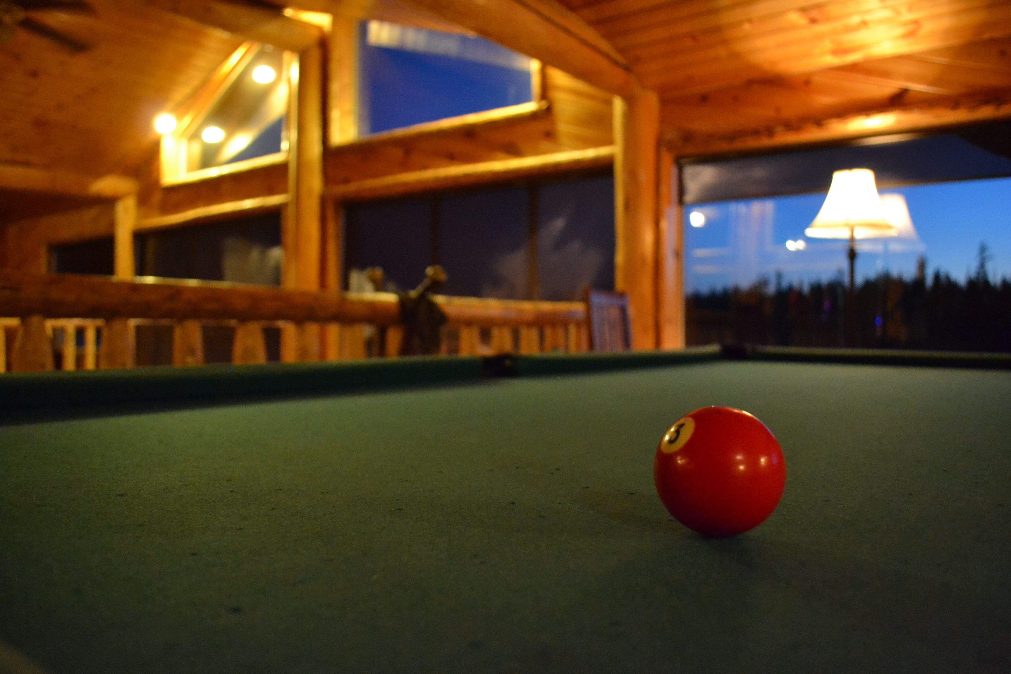 A pool table with a red ball in the foreground, wooden walls, and a window with a lamp outside.