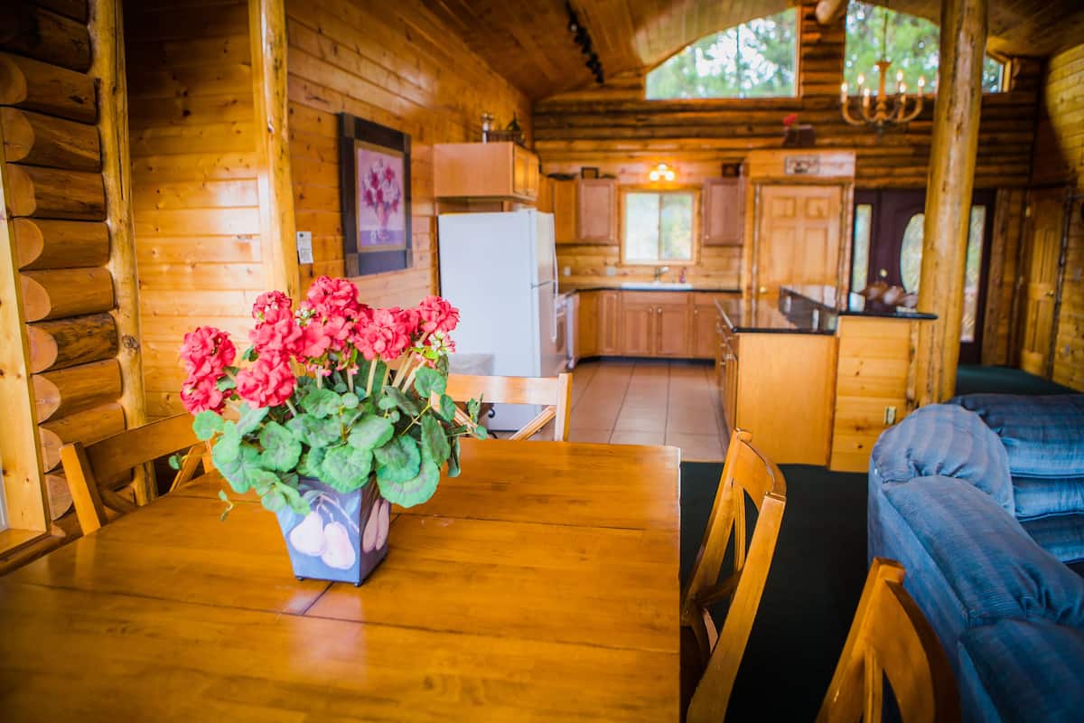 A wooden cabin interior with a dining table, chairs, a sofa, and a kitchen area.
