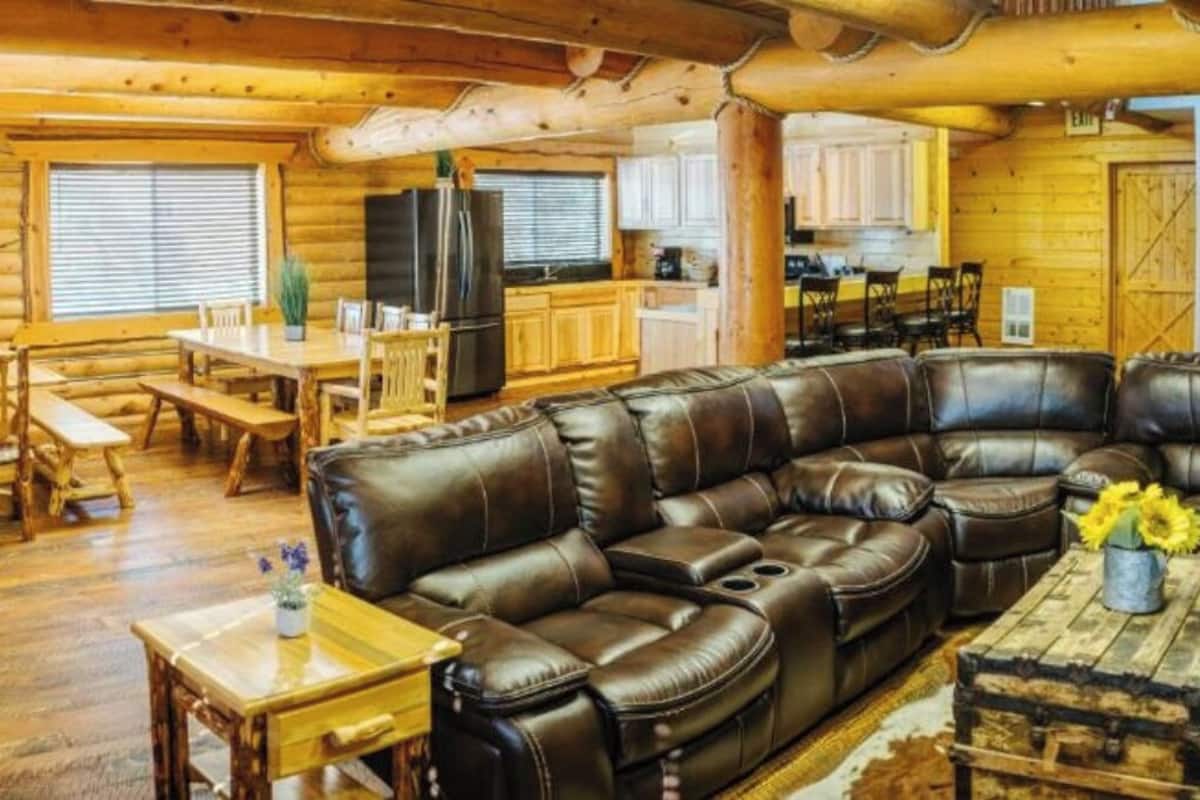 A log cabin interior with a leather sectional sofa, wooden dining table, and a kitchen area.