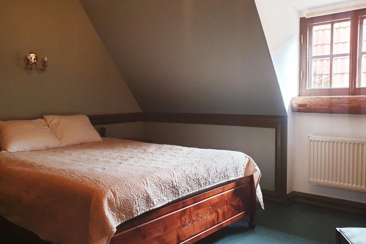 Quarto com teto inclinado, cama de madeira com cabeceira, radiador e janela com cortinas.