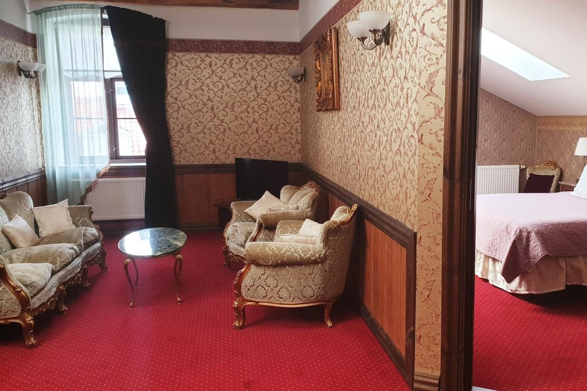 Quarto de hotel com carpete vermelho, duas poltronas, uma mesa pequena, uma cama com colcha rosa e uma janela grande com cortinas.