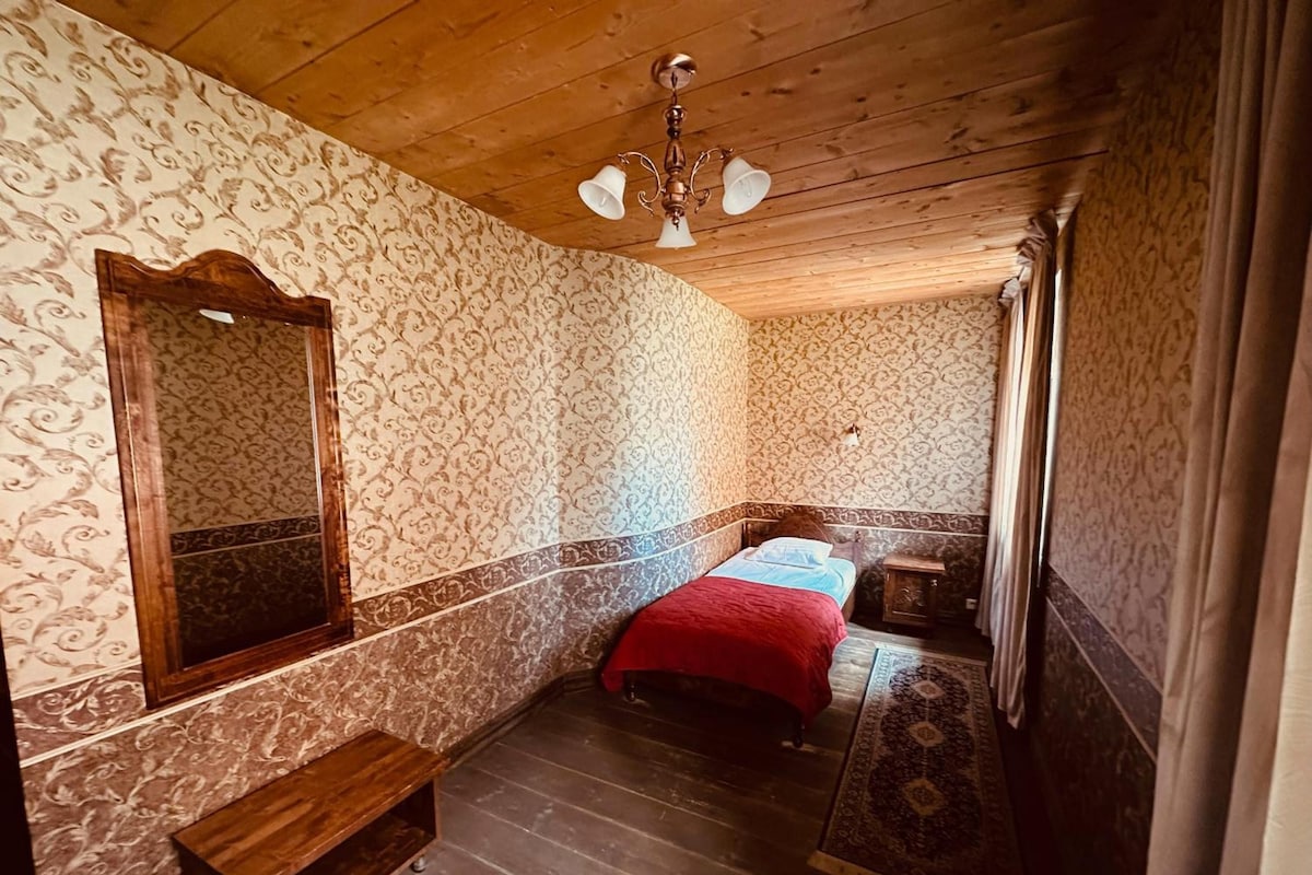 Quarto com teto de madeira, papel de parede estampado, cama com cobertor vermelho, um banco e um pequeno armário.