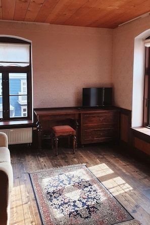 Una habitación con techo de madera, un ventanal grande, televisor, escritorio, silla, radiador y una alfombra estampada.