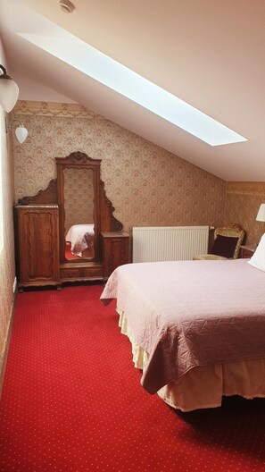 Quarto com teto inclinado, guarda-roupa de madeira, radiador, cama com tapete vermelho e papel de parede estampado.