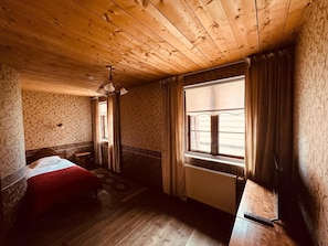 Quarto com teto de madeira, cama com manta vermelha, janela com cortinas e radiador.