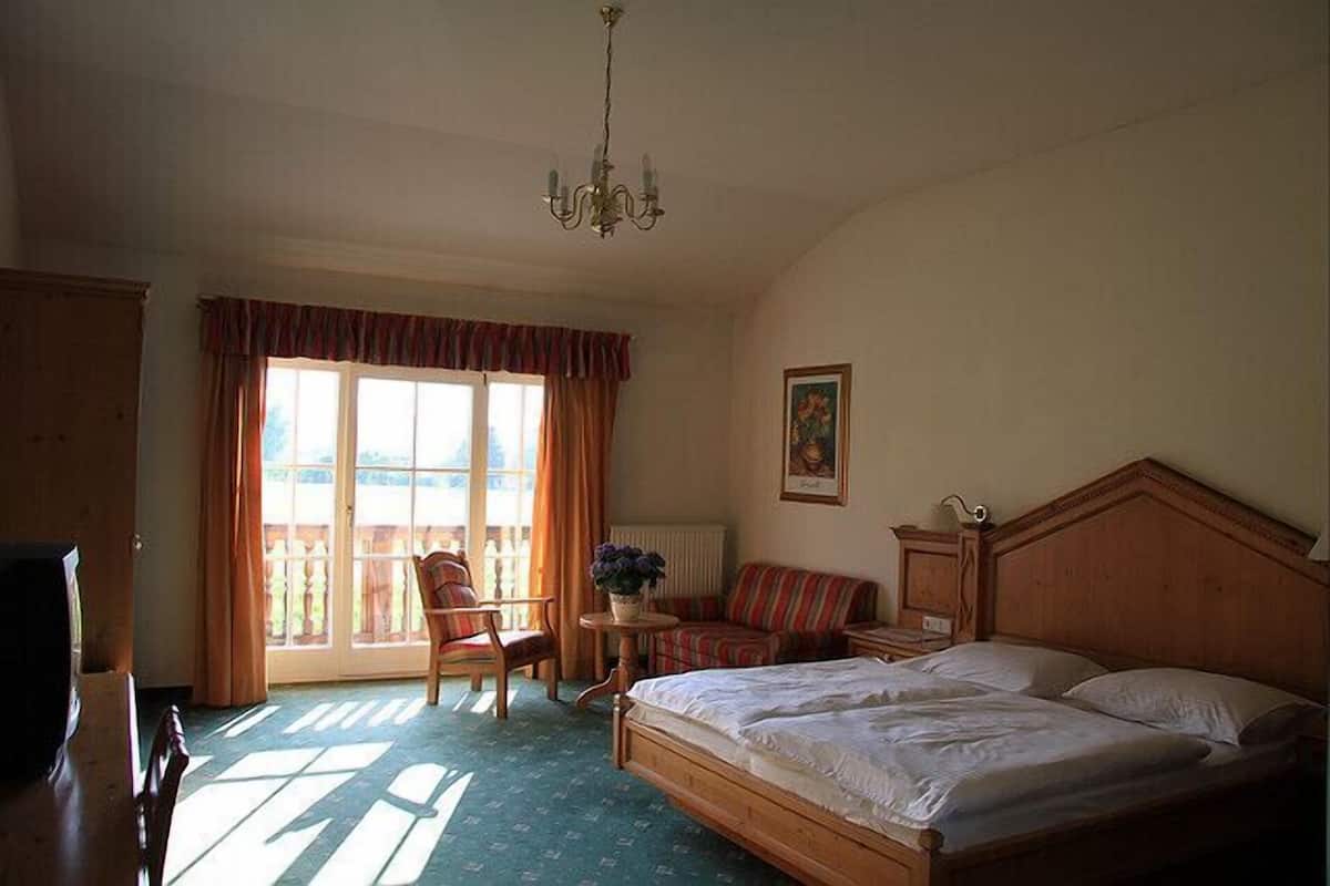 Ein Schlafzimmer mit einem großen Bett, einem Sessel, einem kleinen Tisch mit Pflanze und Blick durch ein fenster mit Vorhängen.