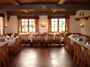 A dining area with wooden tables and chairs, large windows with curtains, and wall-mounted light fixtures. - Hotel Gasthof Mostwastl (Salzburg)
