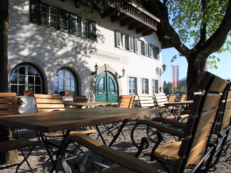 Een terras met houten banken en tafels, een gebouw met gebogen ramen en een groene deur, en een boom die schaduw geeft.