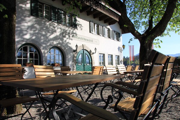 An outdoor seating area with wooden benches and tables, a building with arched windows and a green door, and a tree providing shade. - Hotel Gasthof Mostwastl (Salzburg)