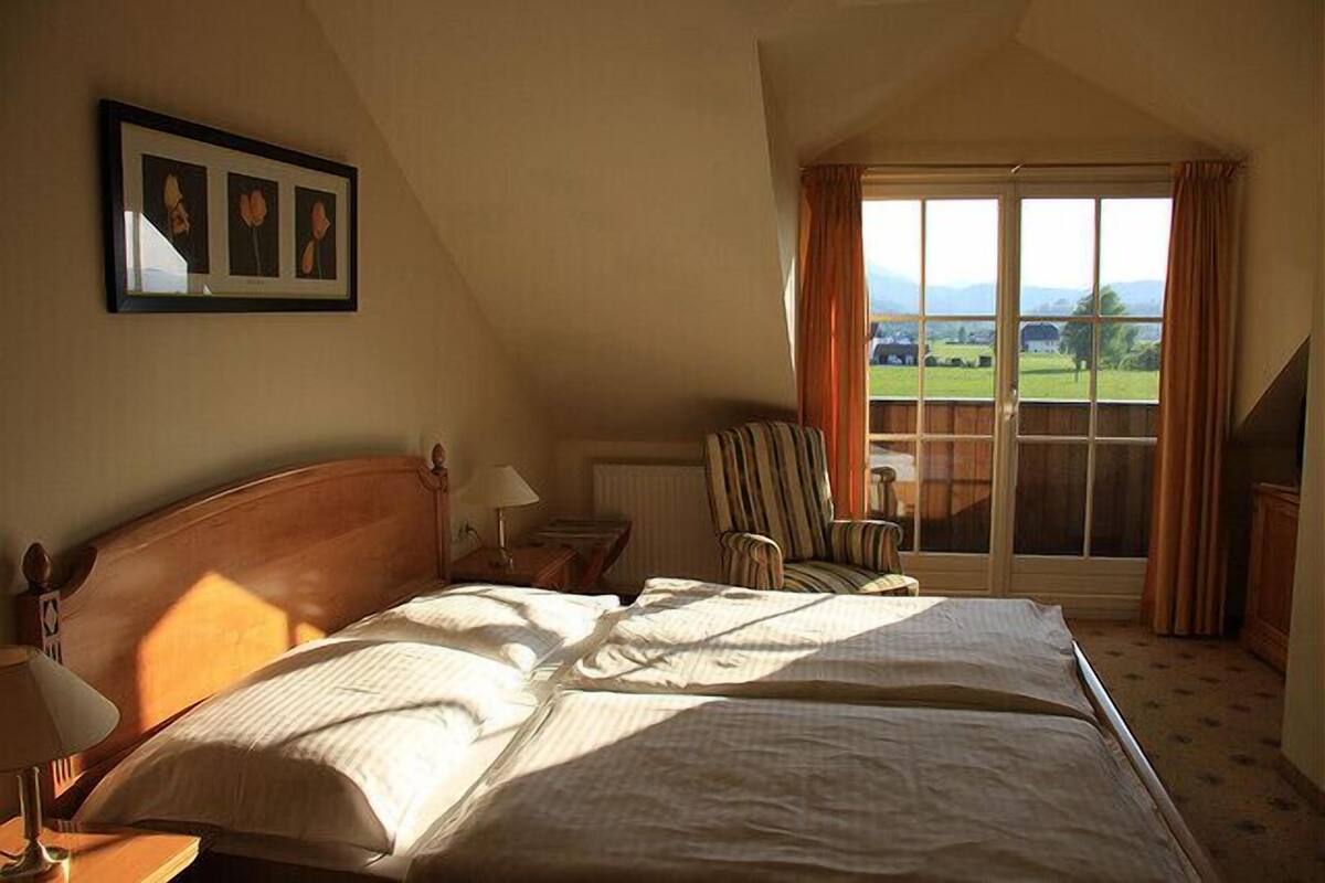 Ein Schlafzimmer mit einem großen Bett, einem Sessel, einem Fenster mit Vorhängen und einem gerahmten Bild an der Wand.