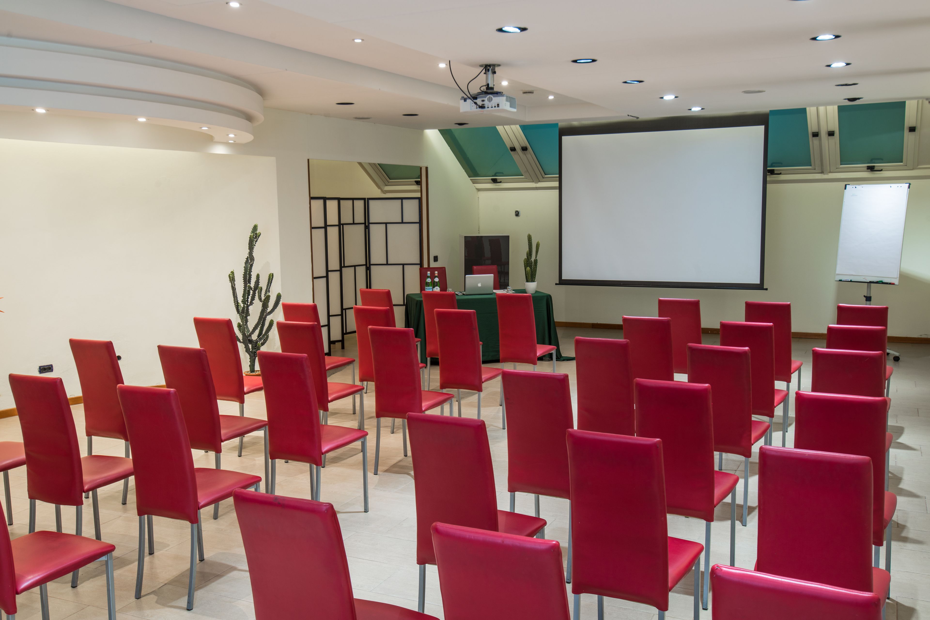 A conference room with red chairs arranged in rows, a projector screen, and a laptop on a table.