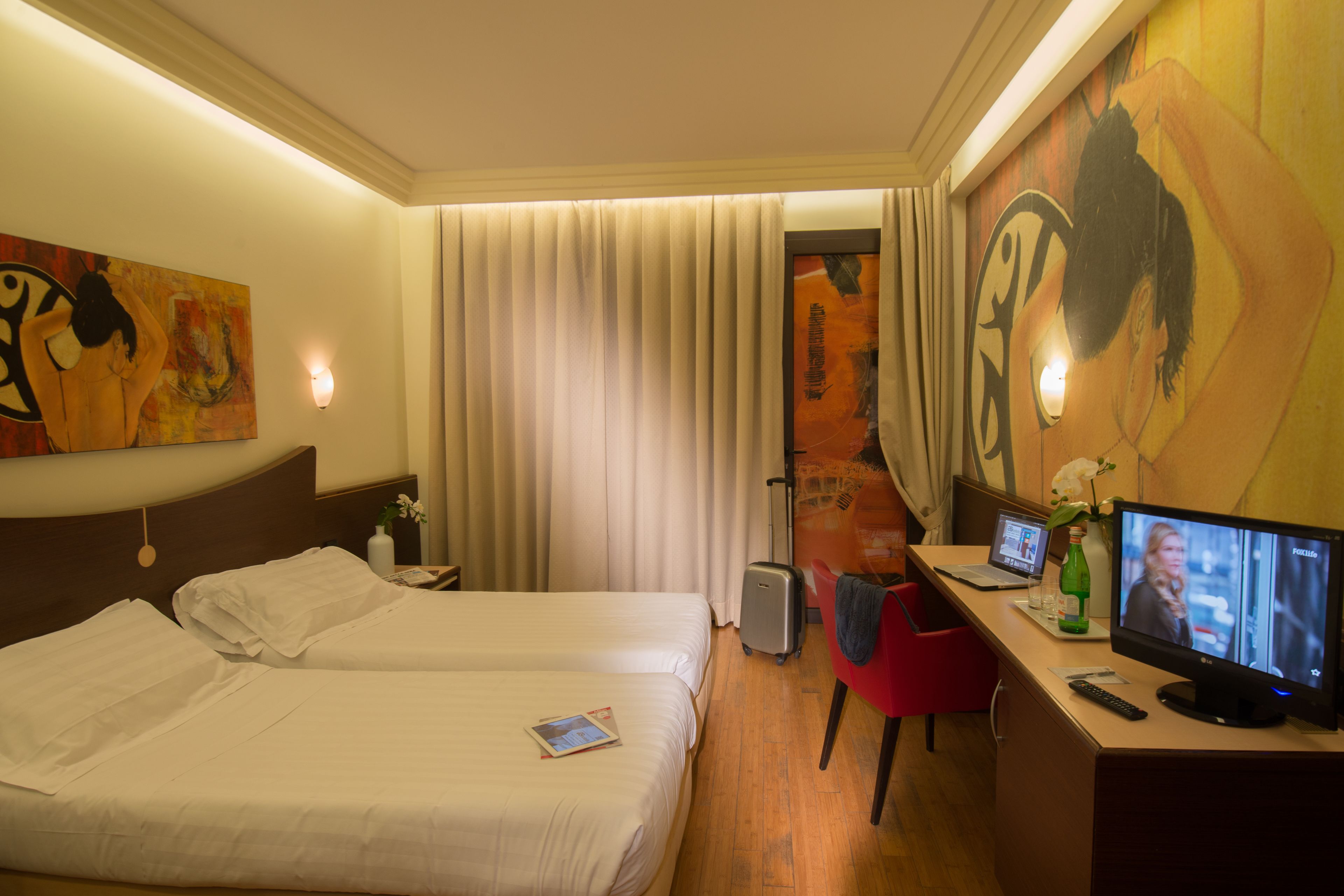 Una habitación de hotel con dos camas, un escritorio con ordenador, un televisor y obras de arte en las paredes.