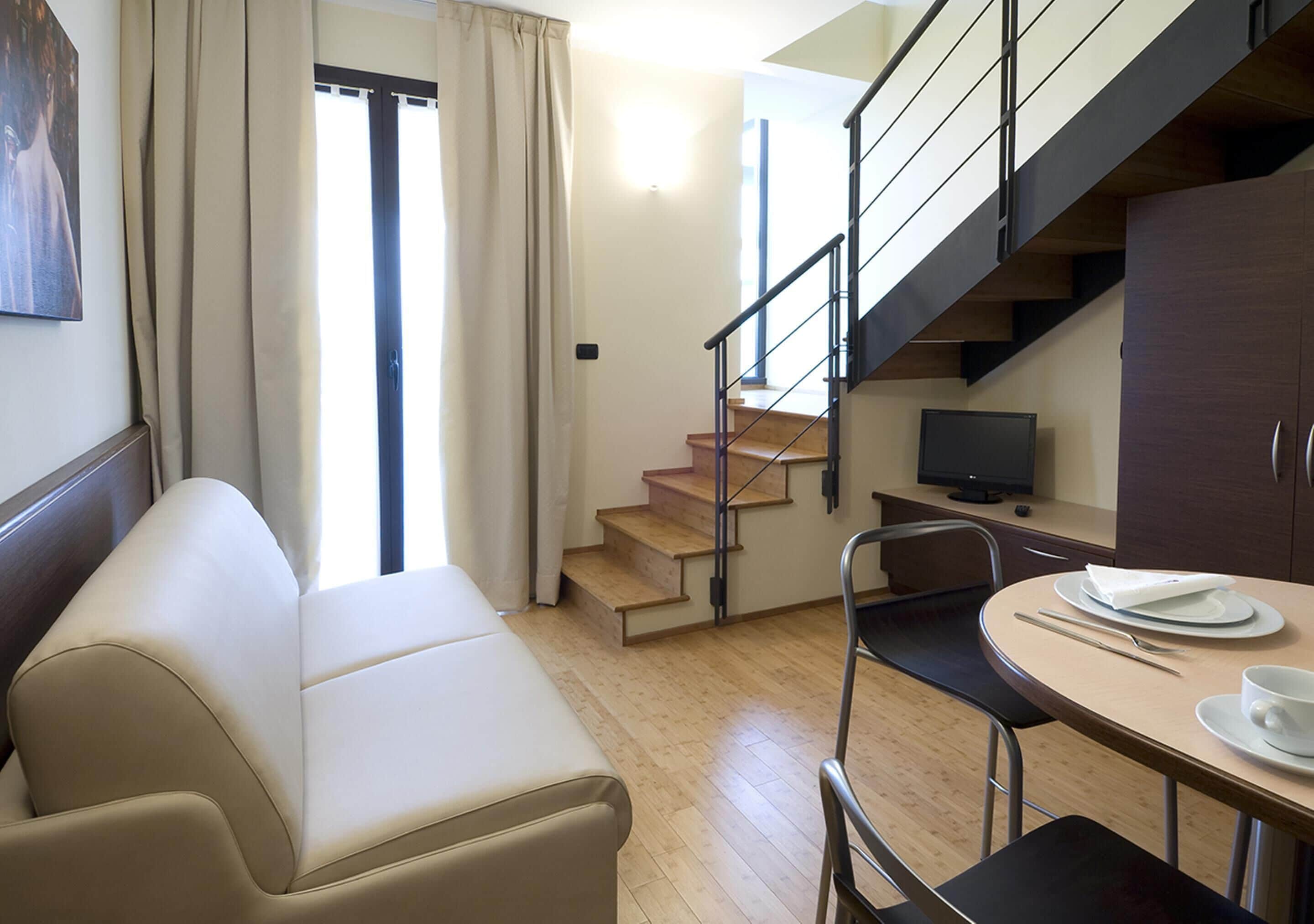 A modern living room with a sofa, a dining table with chairs, a TV, and a staircase.