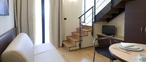 A modern living room with a sofa, a dining table with chairs, a TV, and a staircase.