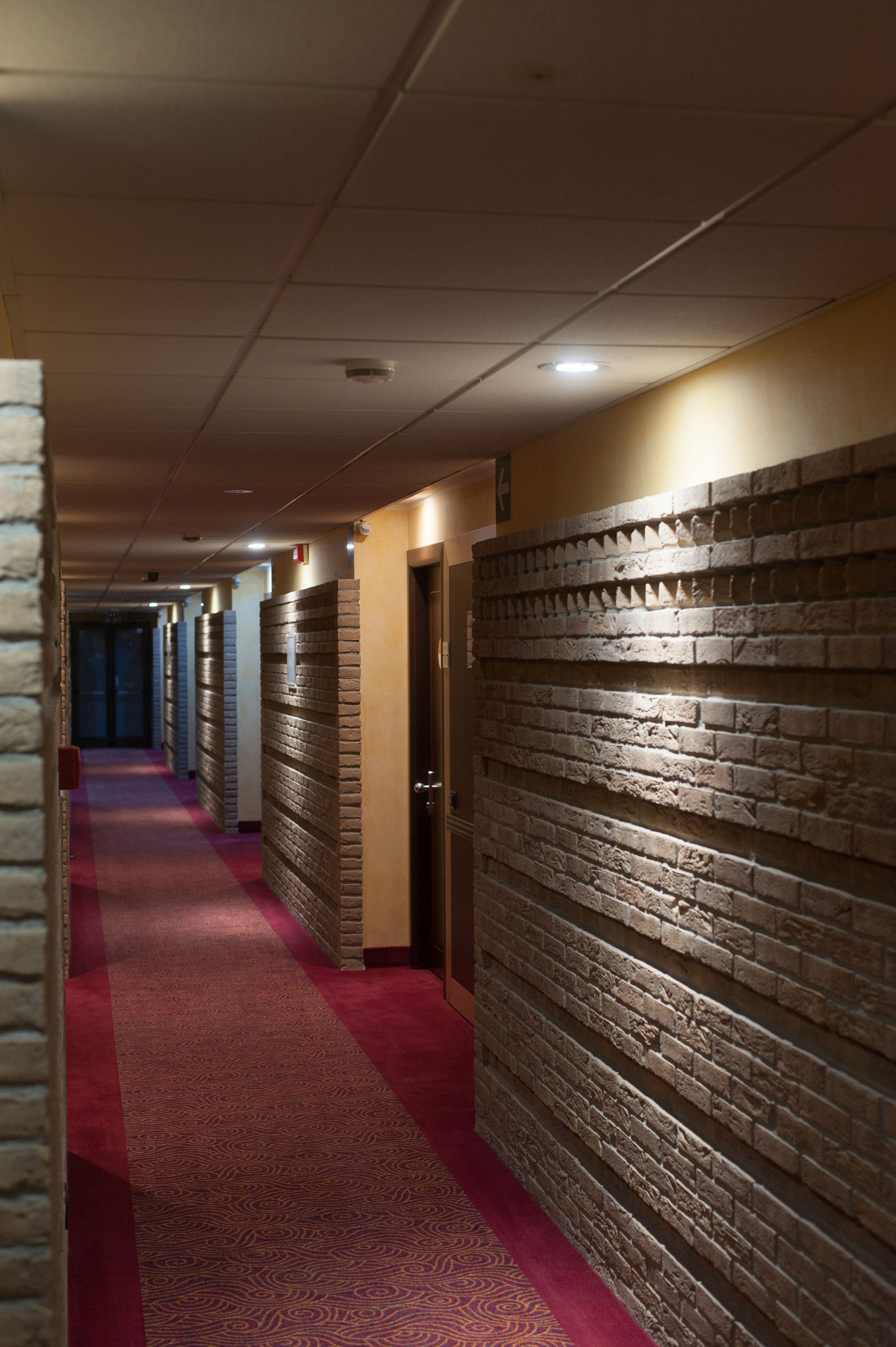 A hotel corridor with a red carpet, brick wall panels, and doors leading to rooms.