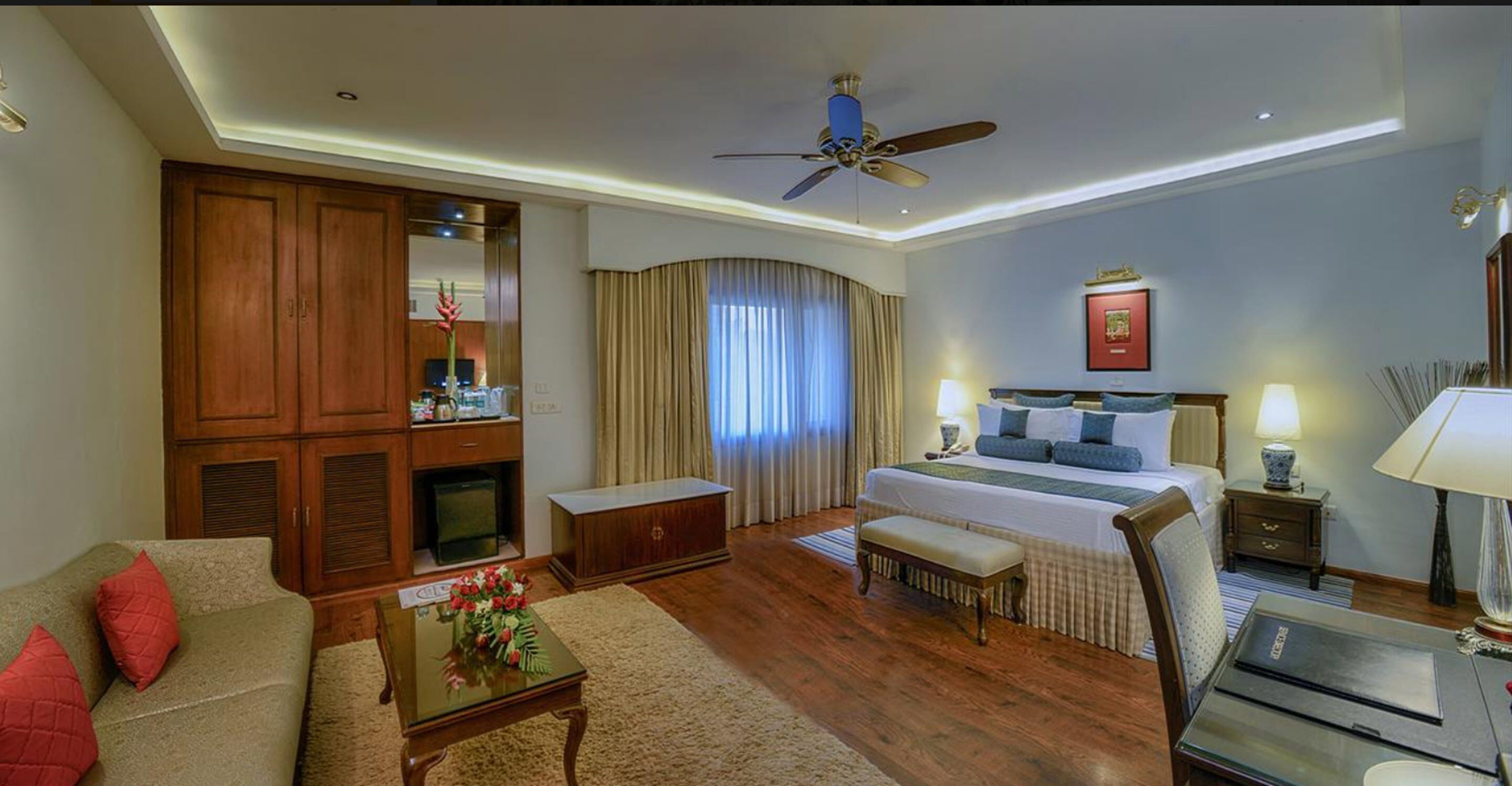 A hotel room with a bed, a sofa, a desk, a wardrobe, and a ceiling fan.