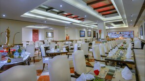 A spacious dining area with multiple tables set for service, a kitchen in the background, and a modern ceiling design. - Hotel Clarks (Varanasi)