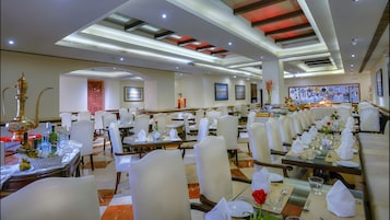 A spacious dining area with multiple tables set for service, a kitchen in the background, and a modern ceiling design.