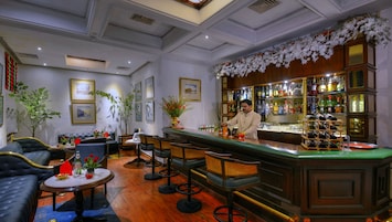 A bar area with a bartender, wooden bar counter, and various bottles. There are seating areas with tables and chairs, and decorative plants.