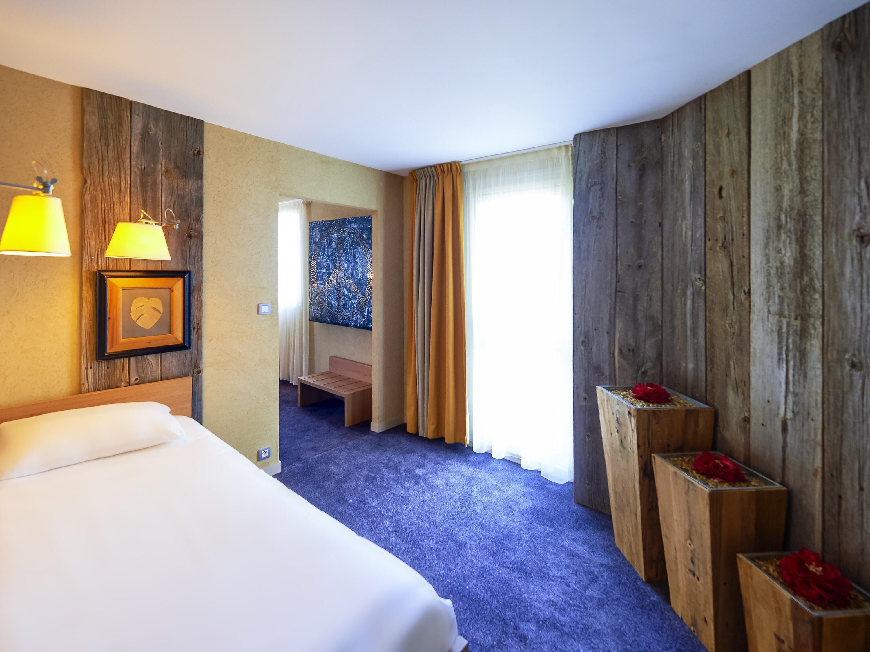 Una habitación de hotel con cama, un banco, pared de madera, un cuadro y una ventana con cortinas.