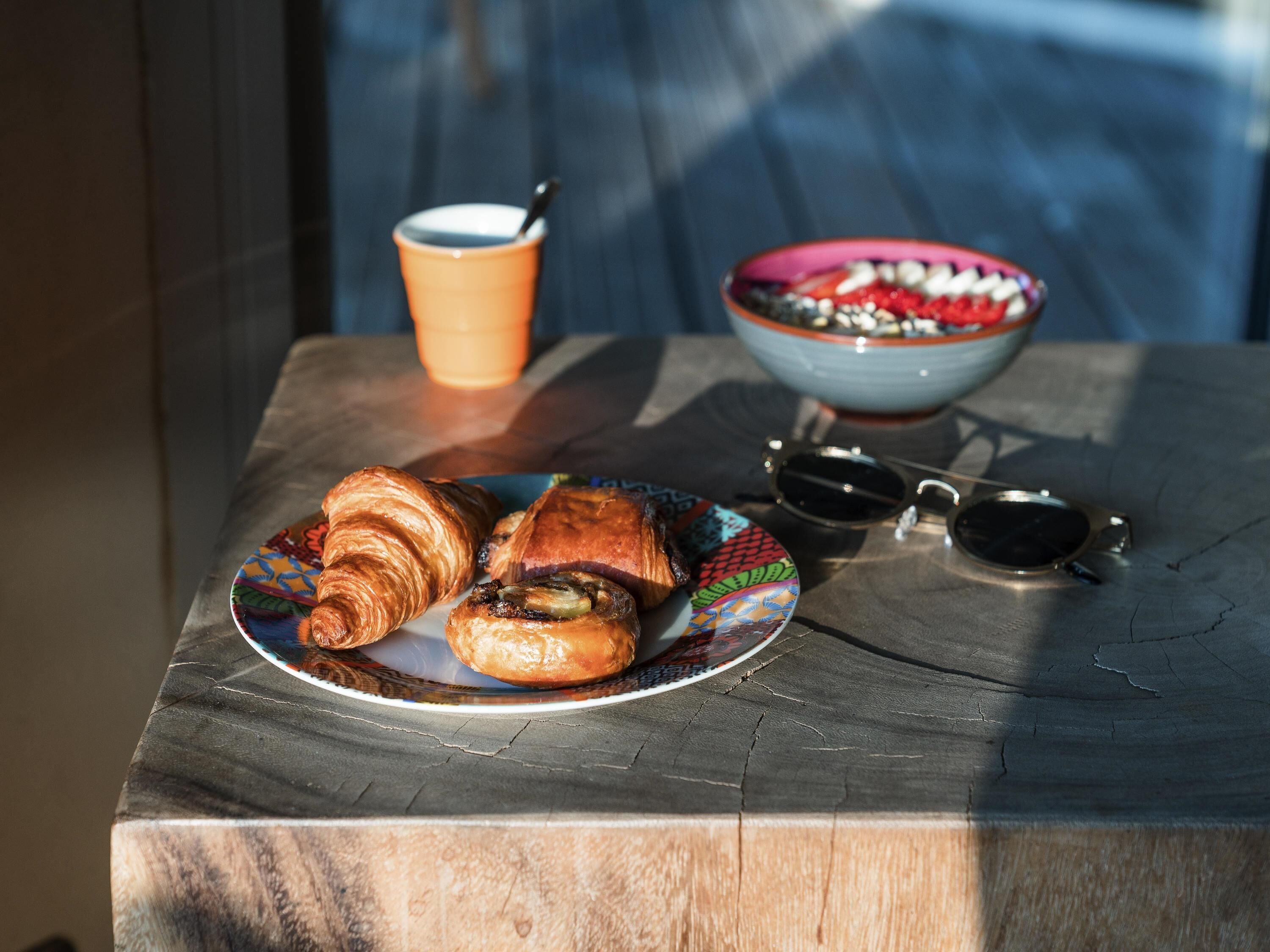 Una mesa con croissants, una taza de café, un bol y gafas de sol.