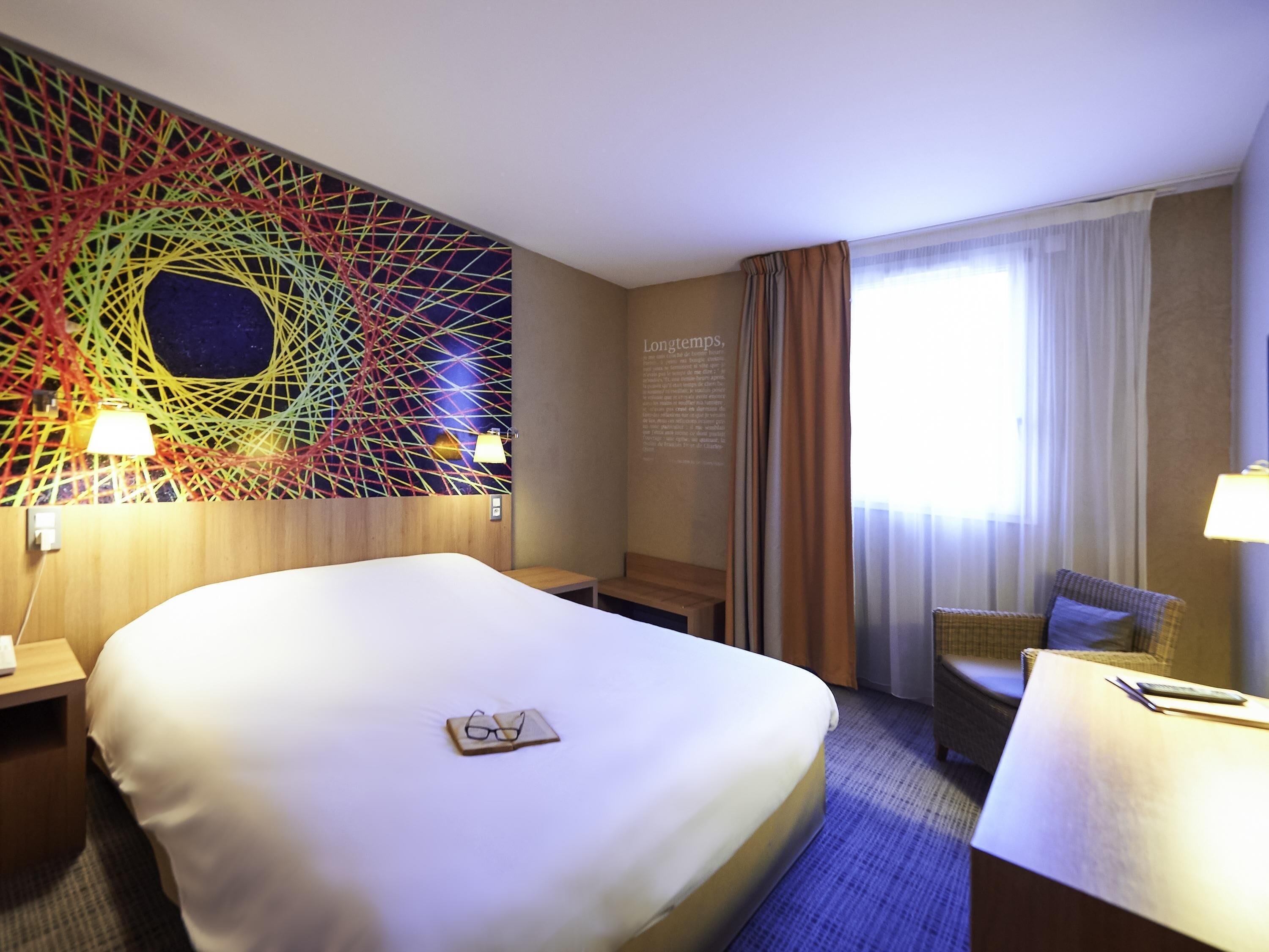 A hotel room with a large bed, a desk, a chair, a wall art piece, and a window with curtains.