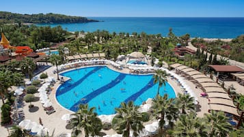 A large outdoor swimming pool surrounded by palm trees and beach umbrellas, with a view of the sea and a distant island.