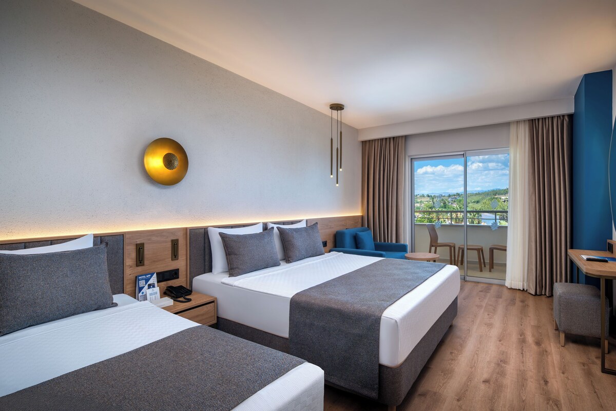 A hotel room with two beds, a desk, a TV, and a balcony with a view.