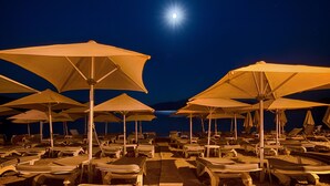 Una playa con filas de tumbonas bajo sombrillas grandes, un pasillo de madera y un mar tranquilo bajo una luna brillante.