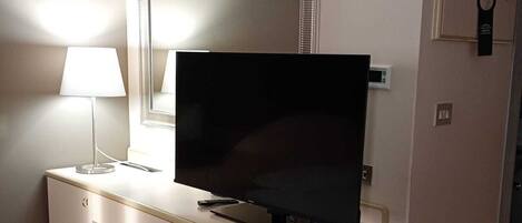 A modern room with a flat-screen TV on a white desk, a lamp, a trash bin, and a mirror on the wall.