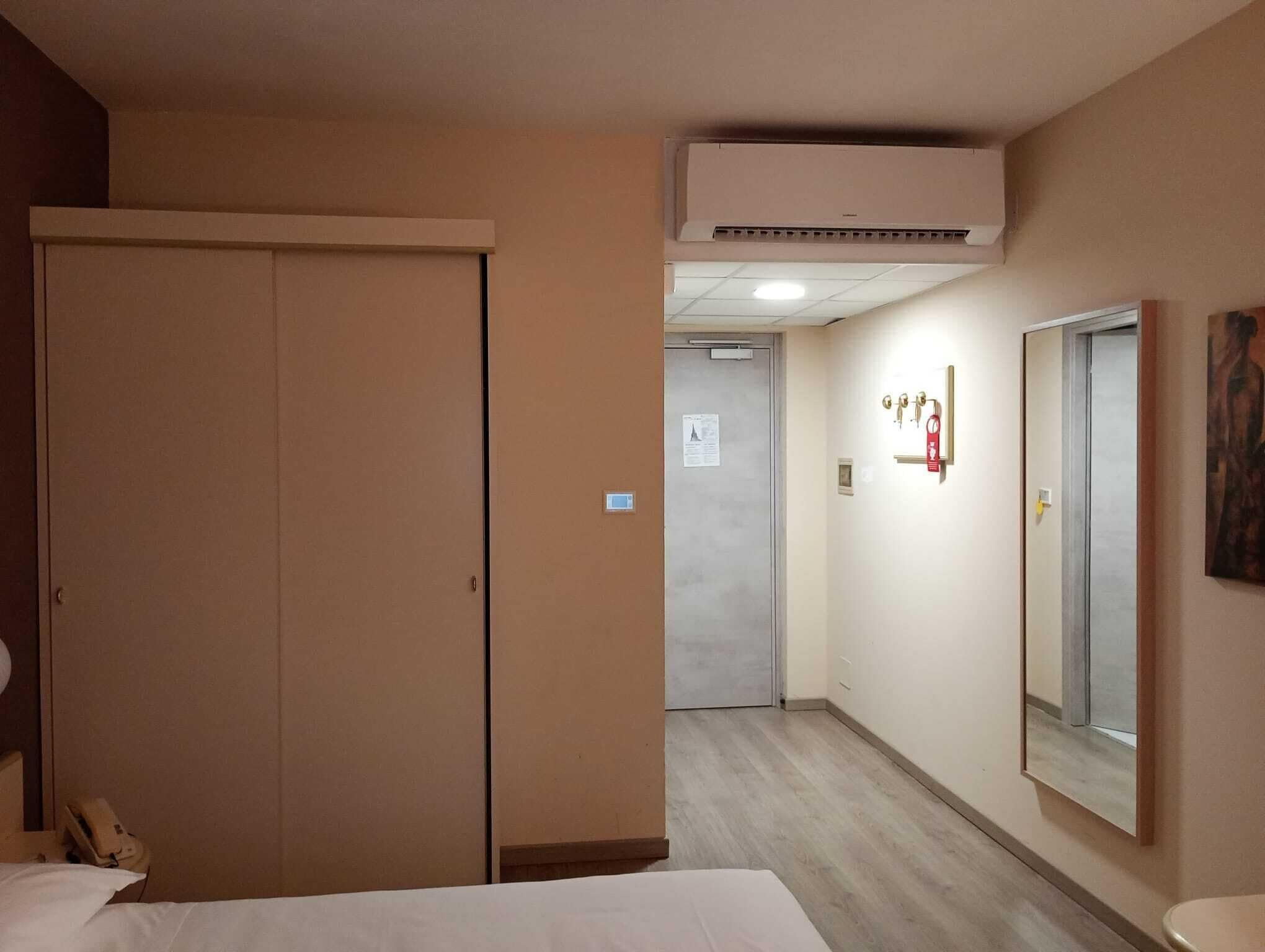 A hotel room with a bed, a wardrobe, a bathroom with a mirror, and an air conditioning unit.