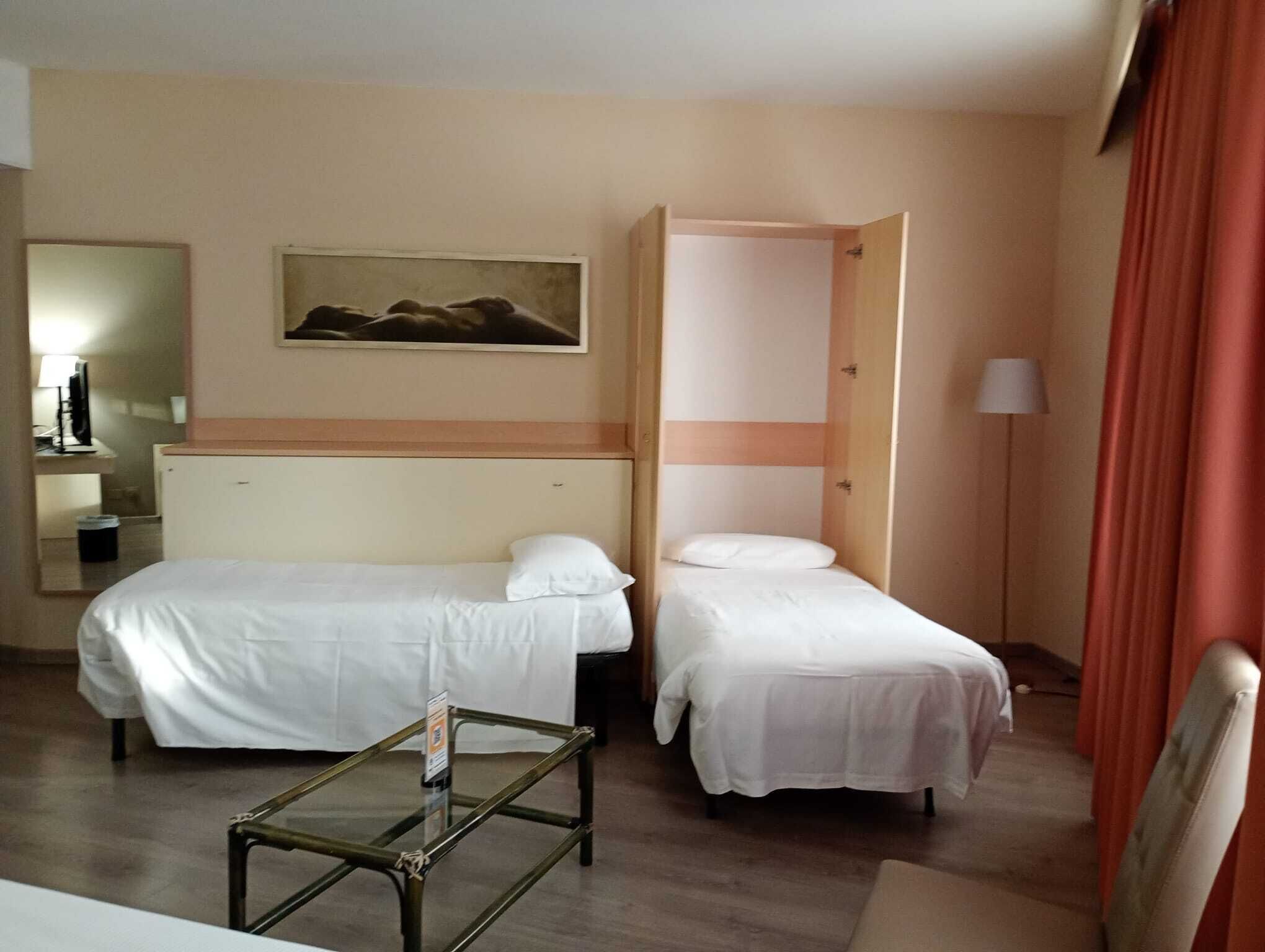 A hotel room with two single beds, a small table, a mirror, a painting on the wall, and a lamp.