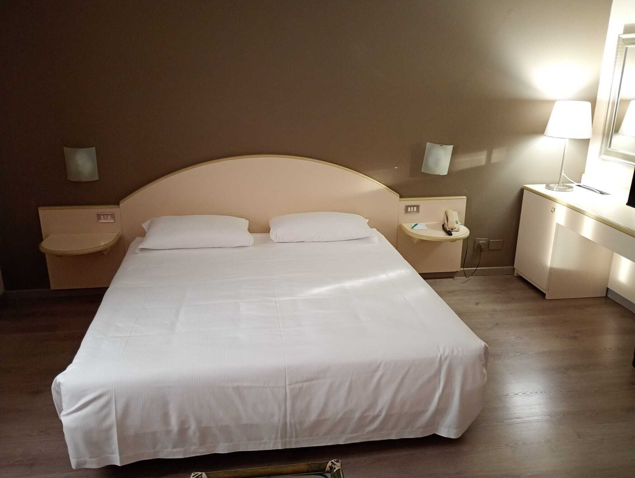 A neatly made bed in a hotel room with a headboard, two bedside tables, a lamp, and a mirror.