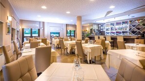 A restaurant with tables set for dining, a bar area with shelves of bottles, and a view of the outdoors through large windows. - Hotel Des Alpes (Rosta)