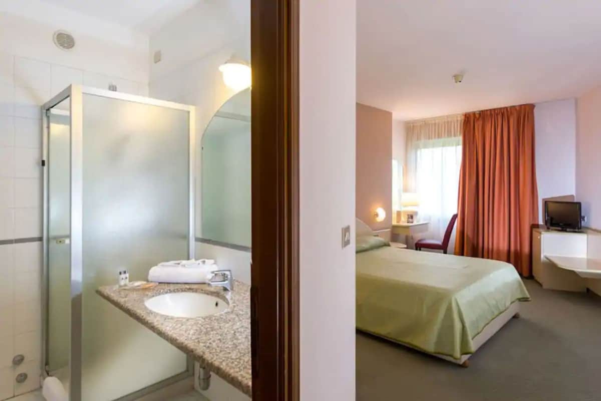 A bathroom with a sink, mirror, and shower. The room has a bed, a desk with a chair, and a TV.
