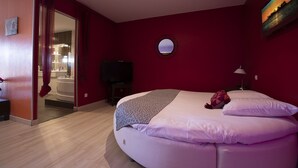 A bedroom with a bed, a round mirror, a television, and a bathroom visible through an open door. -  In Jungle hôtel (Beauvallon)