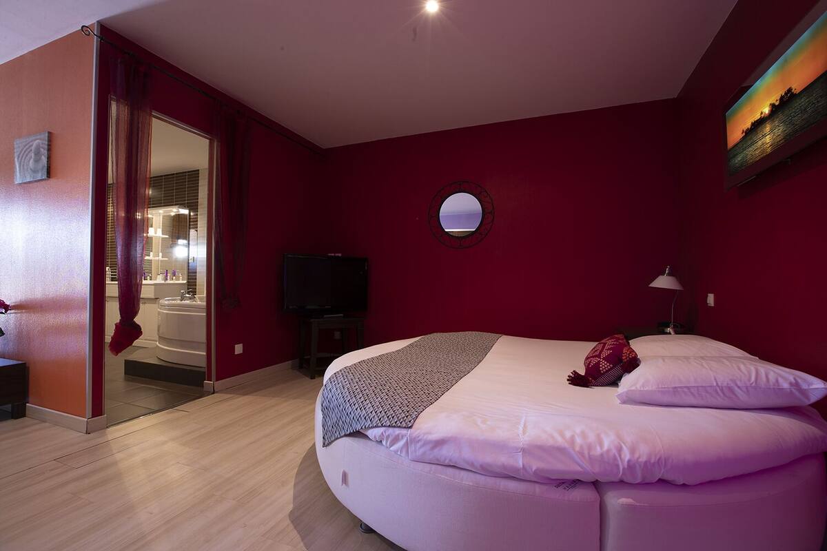 A bedroom with a bed, a round mirror, a television, and a bathroom visible through an open door.