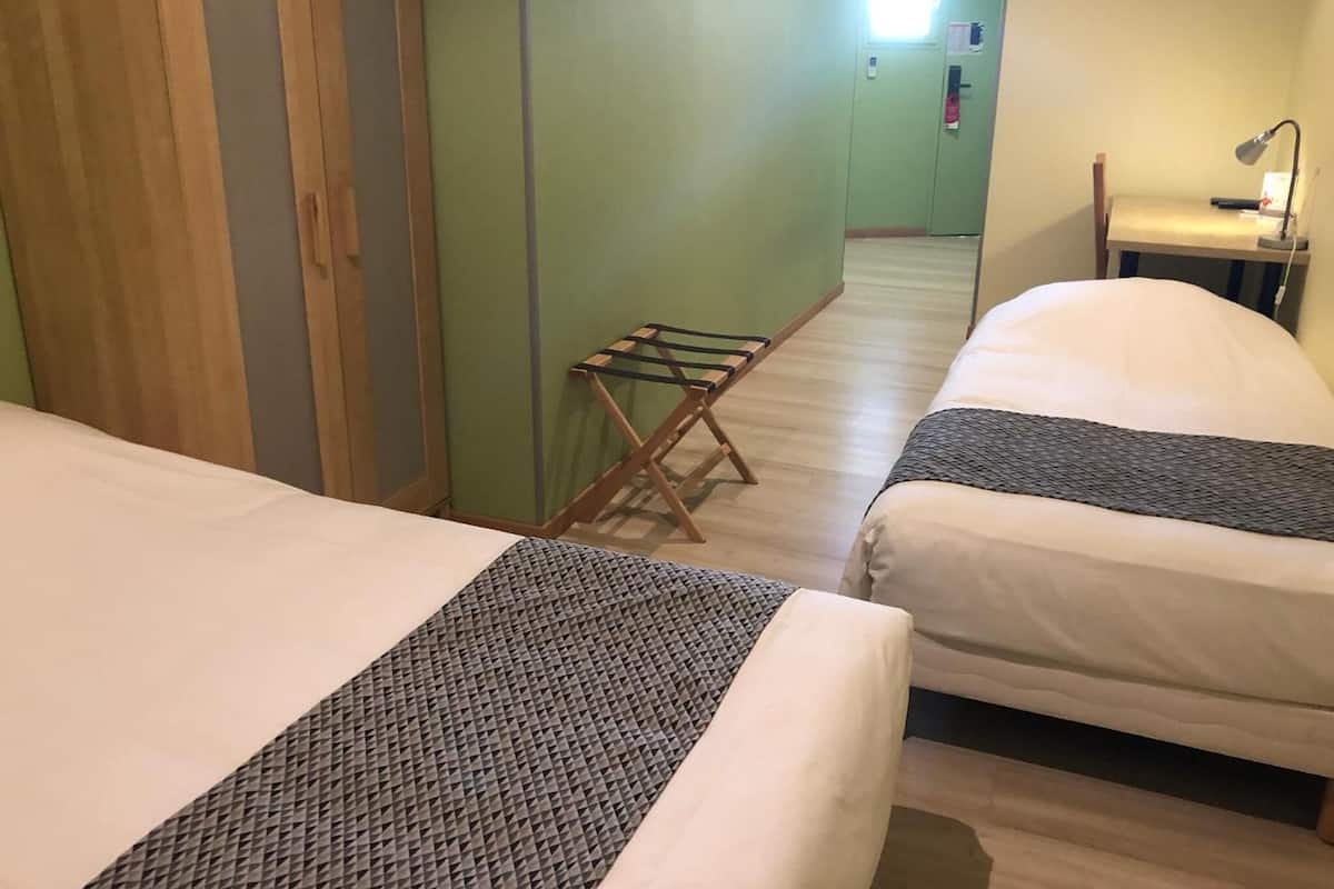 A hotel room with two beds, a wooden wardrobe, a small table with a lamp, and a window with blinds.