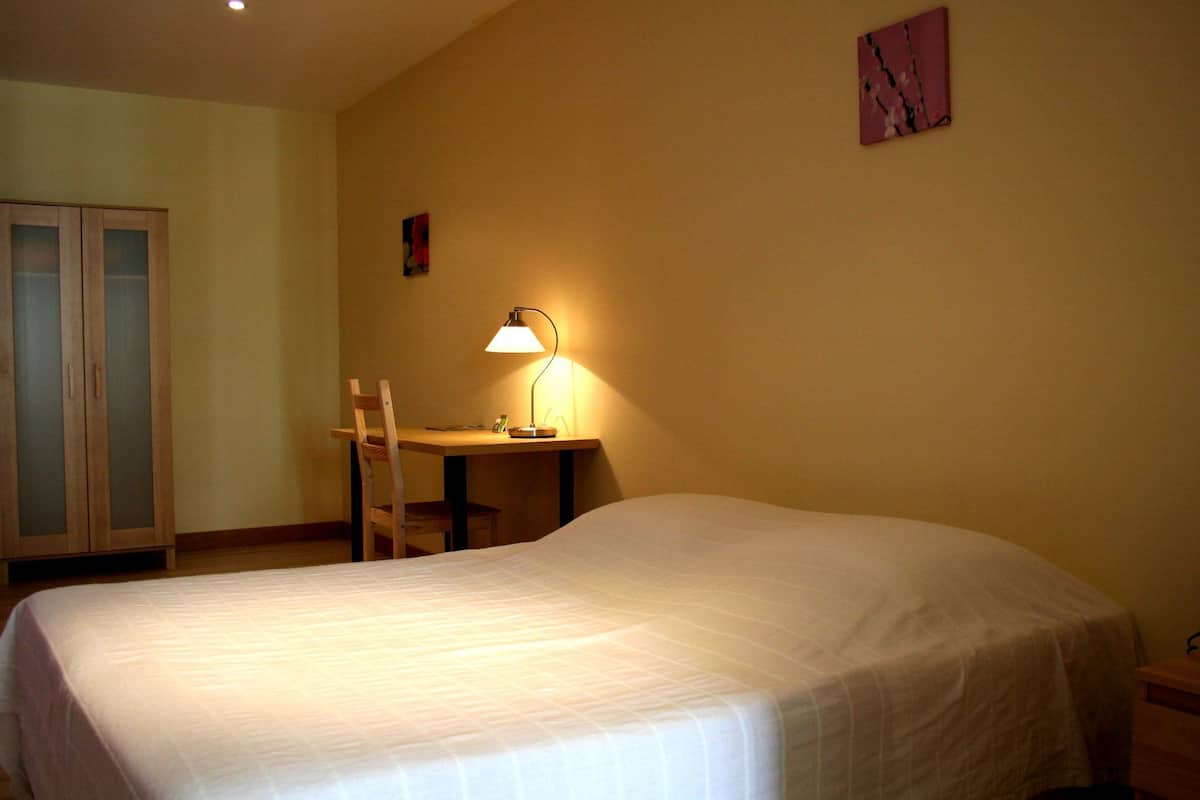 A bedroom with a bed, a desk with a lamp, a chair, and a wardrobe.