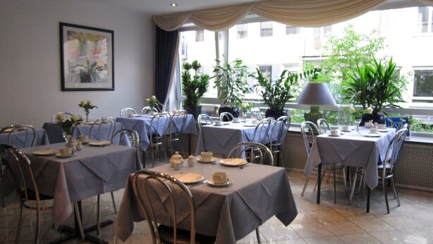 Un espace repas avec des tables dressées, de grandes fenêtres offrant une vue sur les bâtiments et des plantes d’intérieur.
