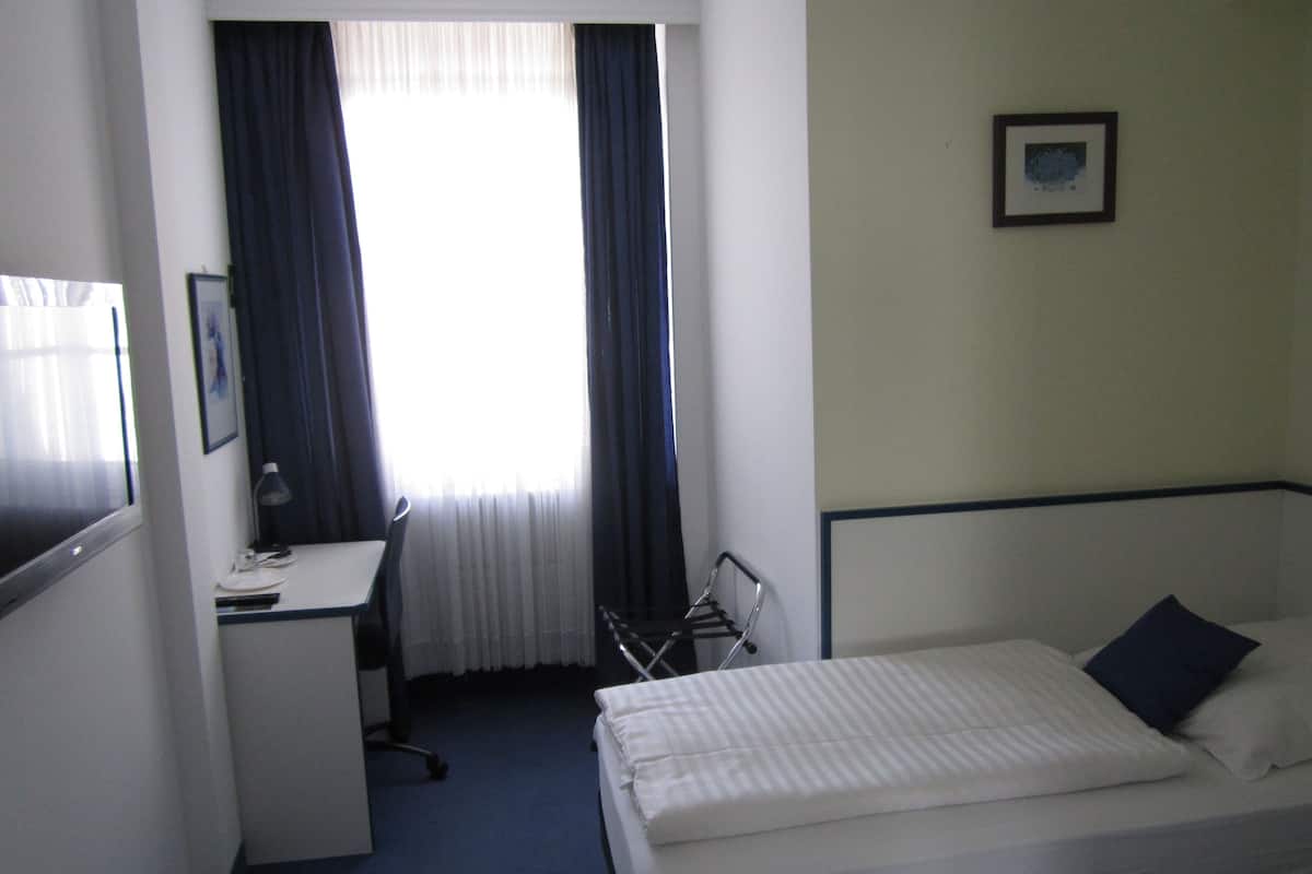 Ein Hotelzimmer mit Bett, Schreibtisch und Stuhl. Es gibt ein Fenster mit Vorhängen, ein Bild an der Wand und einen kleinen Tisch mit Lampe.