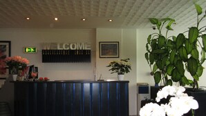 Un comptoir d’accueil avec une pancarte « BIENVENUE », un tableau encadré accroché au mur et une plante en pot ornée de fleurs blanches.