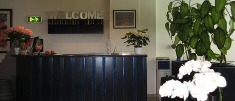 A reception desk with a 'WELCOME' sign, a framed picture on the wall, and a potted plant with white flowers.