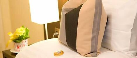 Una cama bien hecha con ropa blanca y dos almohadas decorativas, una mesita de noche con una lámpara, y un pequeño jarrón con flores.