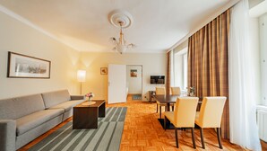 Ett modernt hotellrum med en soffa, ett matbord, stolar, en TV och ett stort fönster med gardiner.