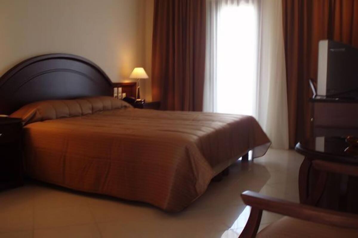 A hotel room with a large bed, a nightstand with a lamp, a desk with a chair, and a television.