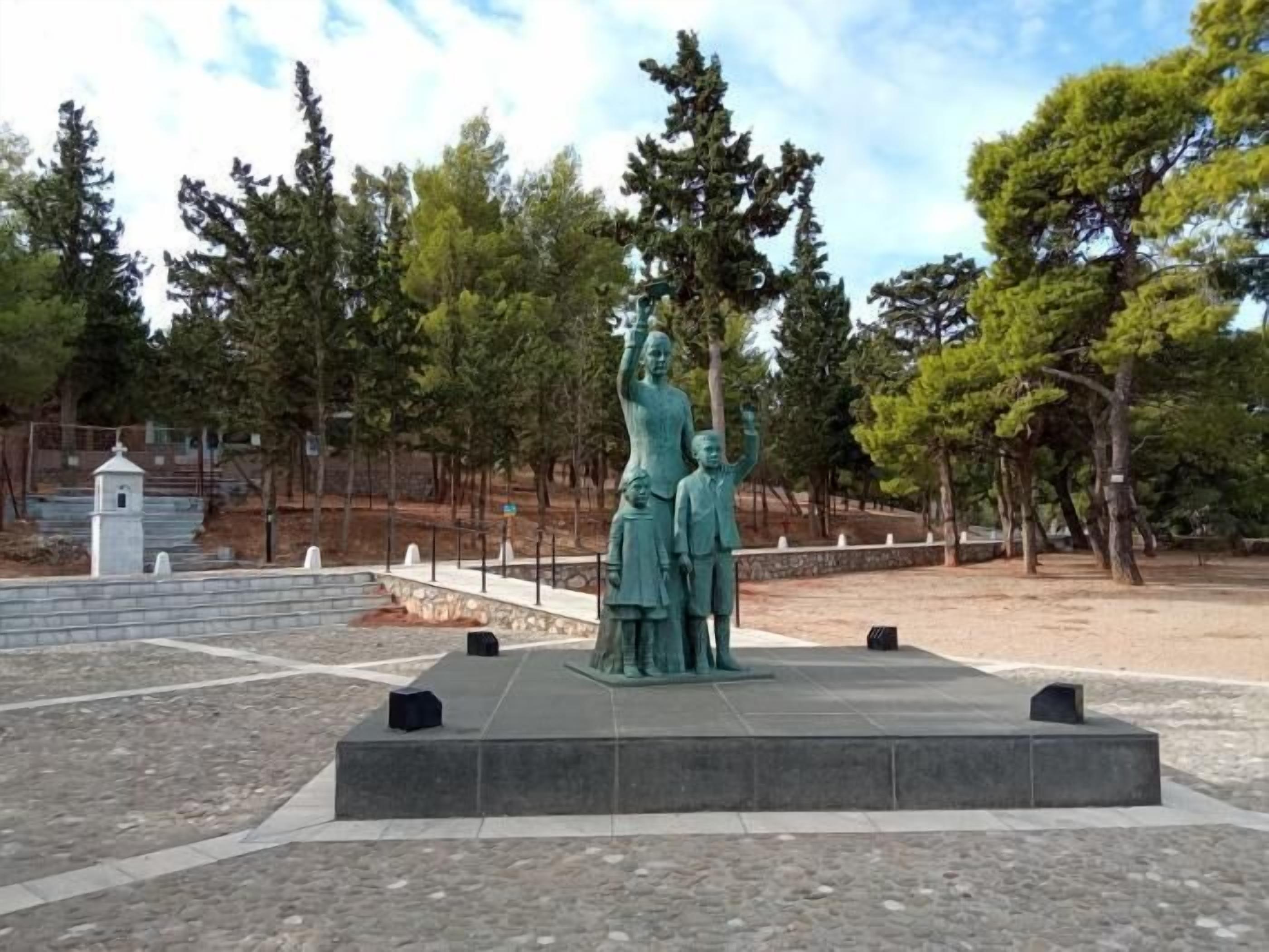 Una estatua en un parque, sobre un pedestal de piedra, rodeada de árboles y un pequeño edificio blanco.