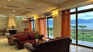 A spacious living area with leather sofas, a fireplace, and large windows offering a view of a lake and mountains. - Europa Beach Hotel (Delphi)