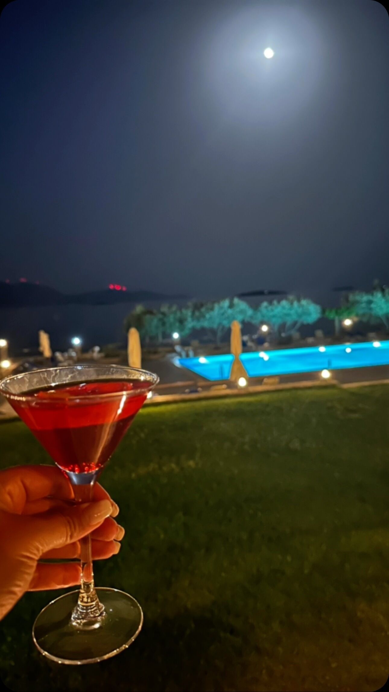 A hand holding a red cocktail glass with a clear liquid, set against a nighttime backdrop featuring a pool and illuminated structures.